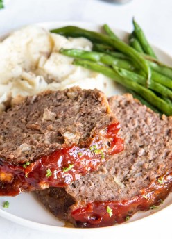 Two slices of meatloaf with glaze served with mashed potatoes and green beans.