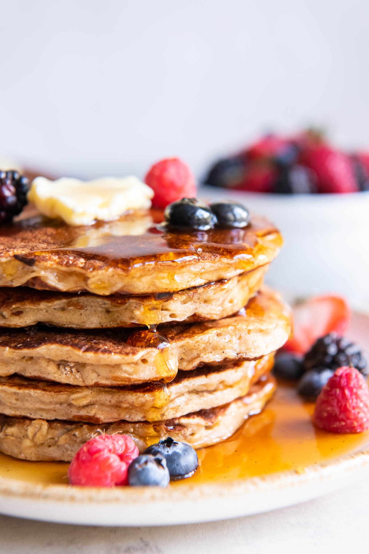 Close up of stack of 5 oatmeal pancakes topped with butter, maple syrup and fresh berries.