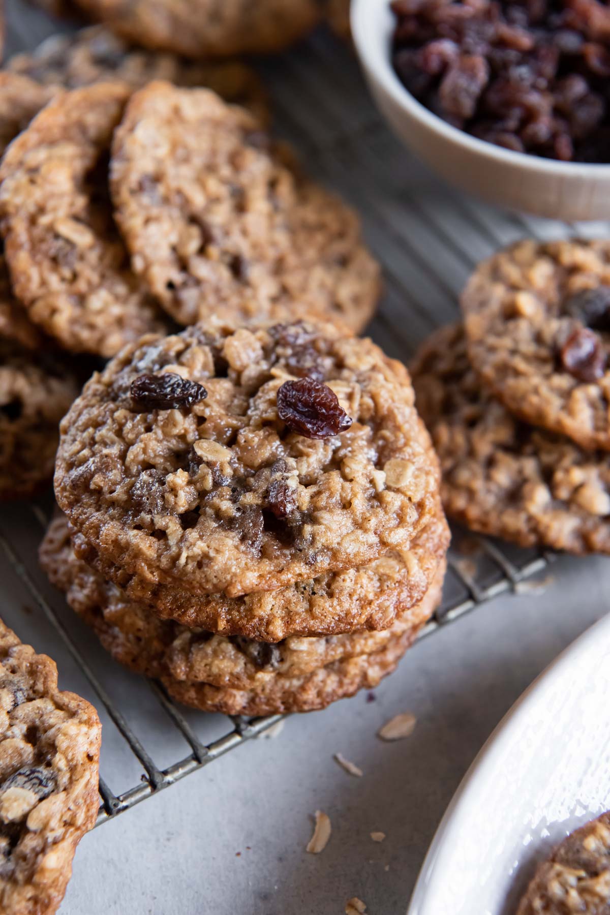 Oatmeal raisin cookies stacked on a cooling rack with a bowl of raisins in the background.