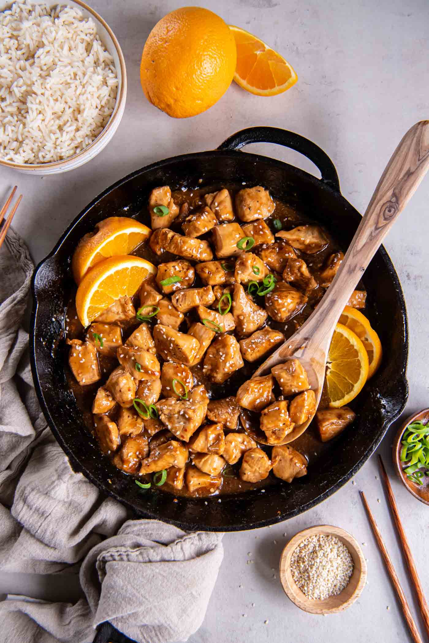 Orange chicken in a cast iron skillet with a wooden spoon with oranges, rice, sesame seeds and green onions around the skillet.