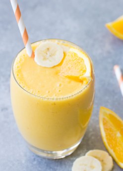 Orange smoothie in a glass with a straw with banana slices and orange wedges.