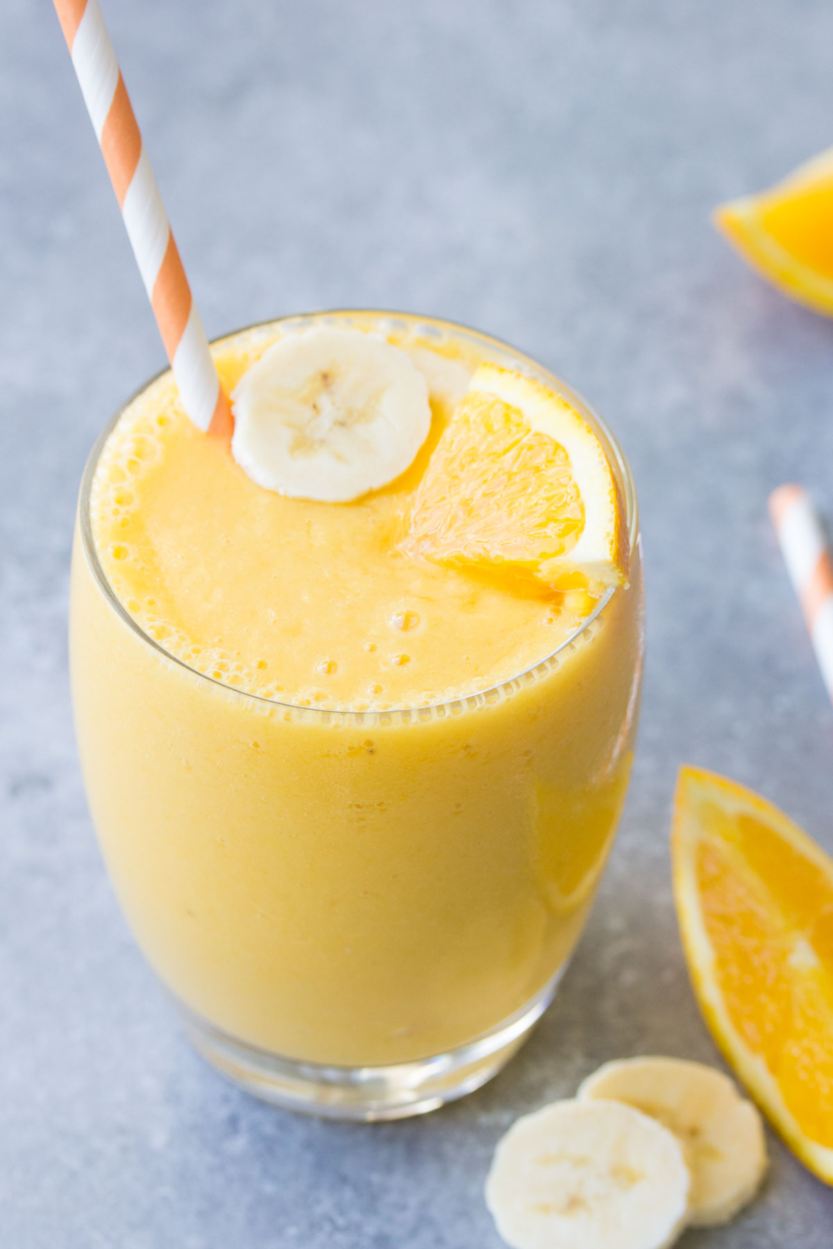 Orange smoothie in a glass with a straw, with banana slice and orange wedge.