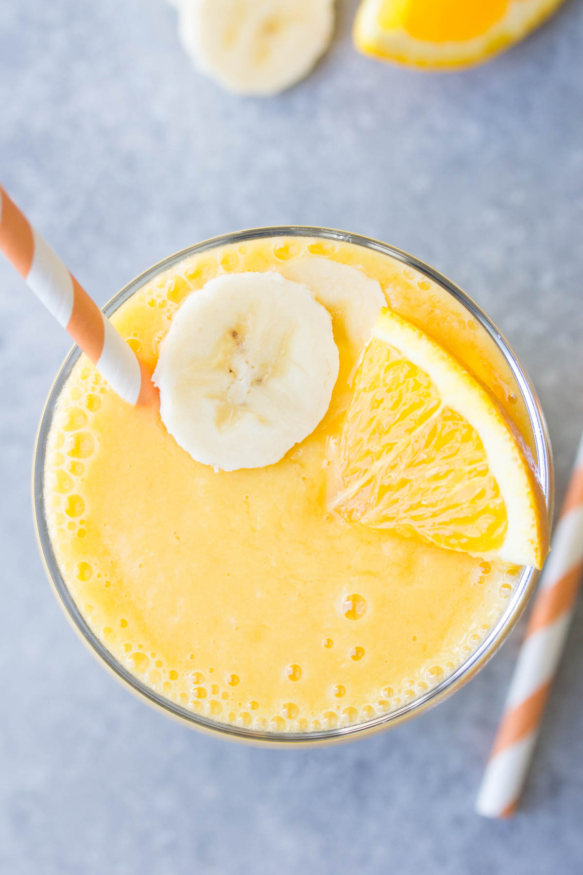 Overhead view of orange smoothie in glass with straw, banana slice and orange wedge.
