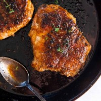 Two pan fried pork chops in a cast iron skillet with a spoon for the pan sauce.