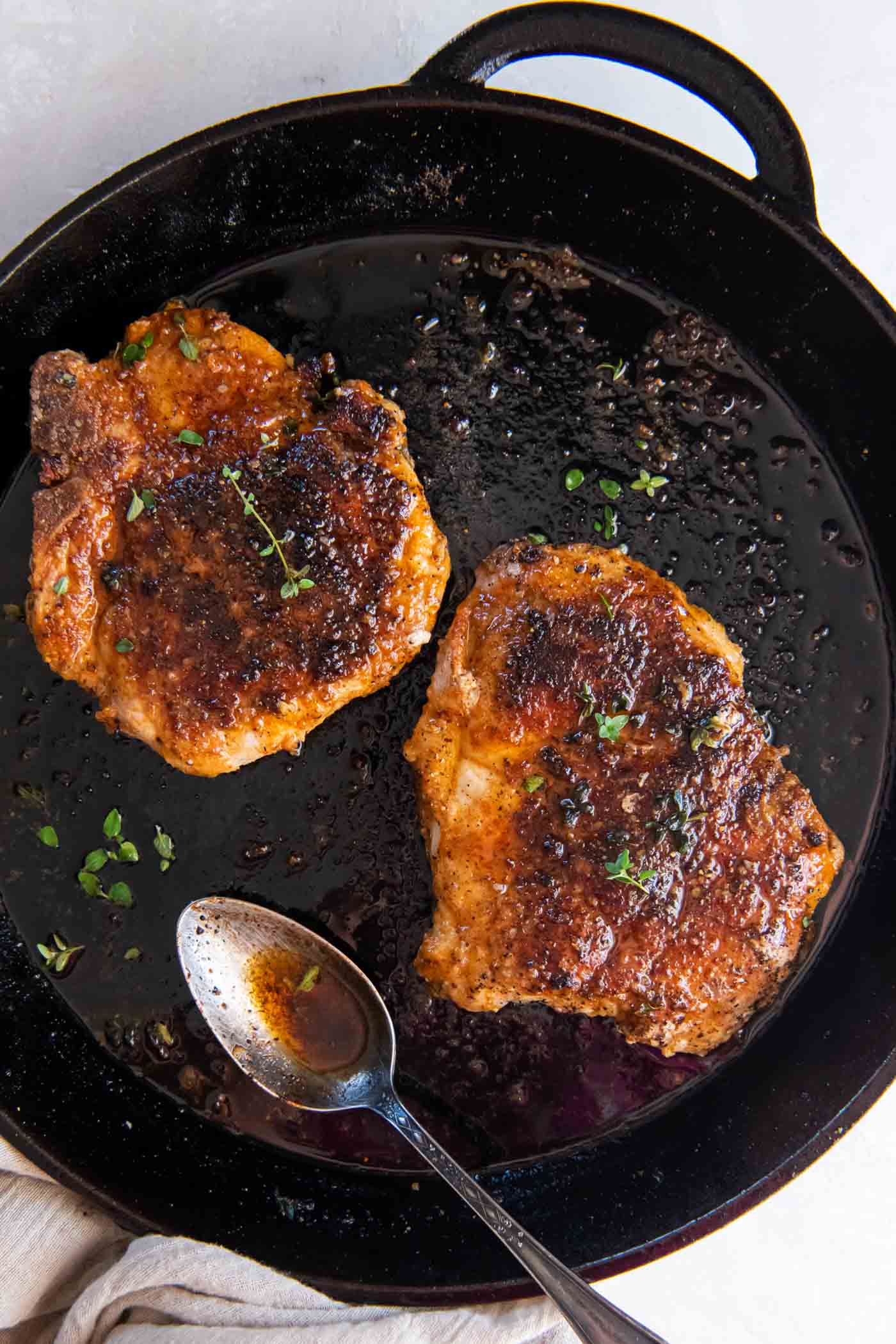 Two pan fried pork chops in a cast iron skillet with a spoon.