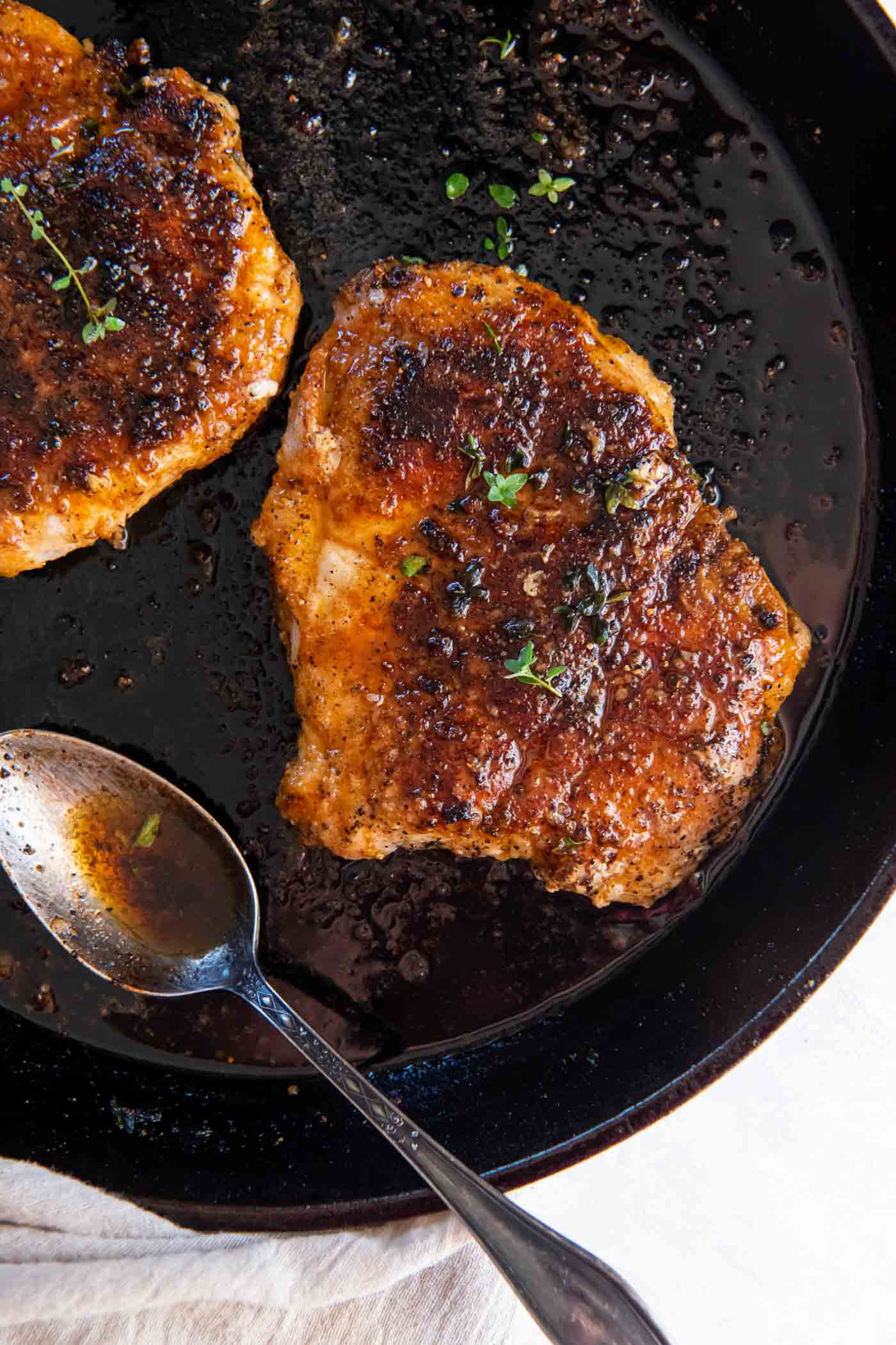Two bone-in pan fried pork chops in a cast iron skillet with a spoon for the sauce.