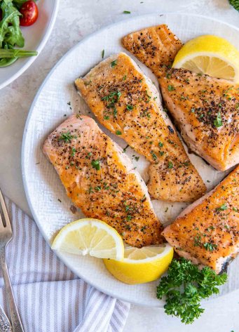 Four pan seared salmon fillets on a plate with lemon wedges.