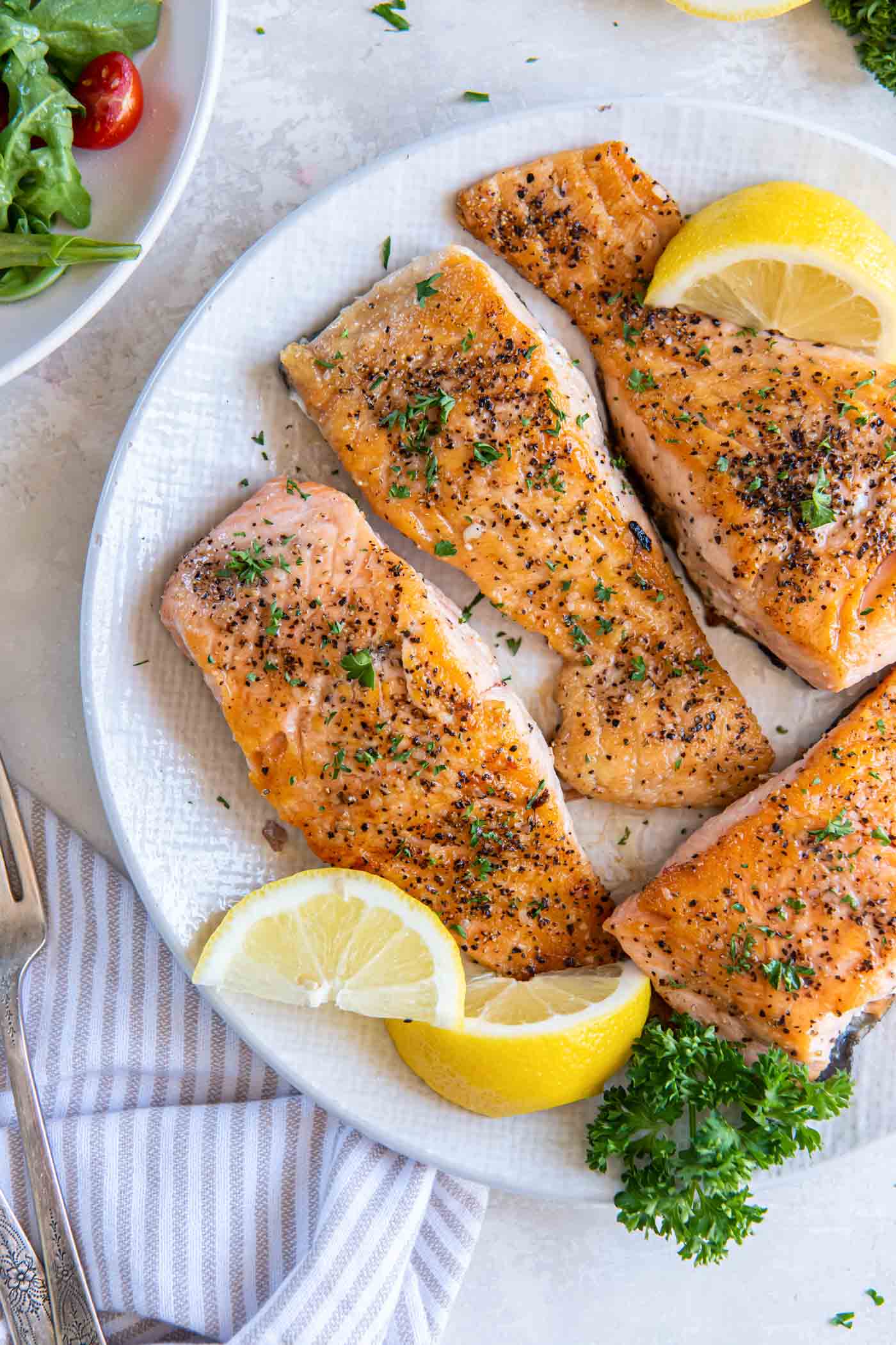 Four pan seared salmon fillets on a plate with lemon wedges.