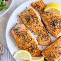Four pan seared salmon fillets on a plate with lemon wedges.
