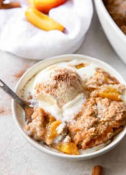 serving of peach cobbler in a bowl topped with a scoop of melty vanilla ice cream and cinnamon