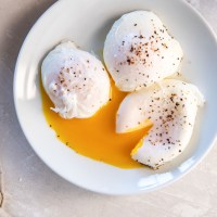 Three poached eggs on a plate with one egg sliced open so yolk runs out.