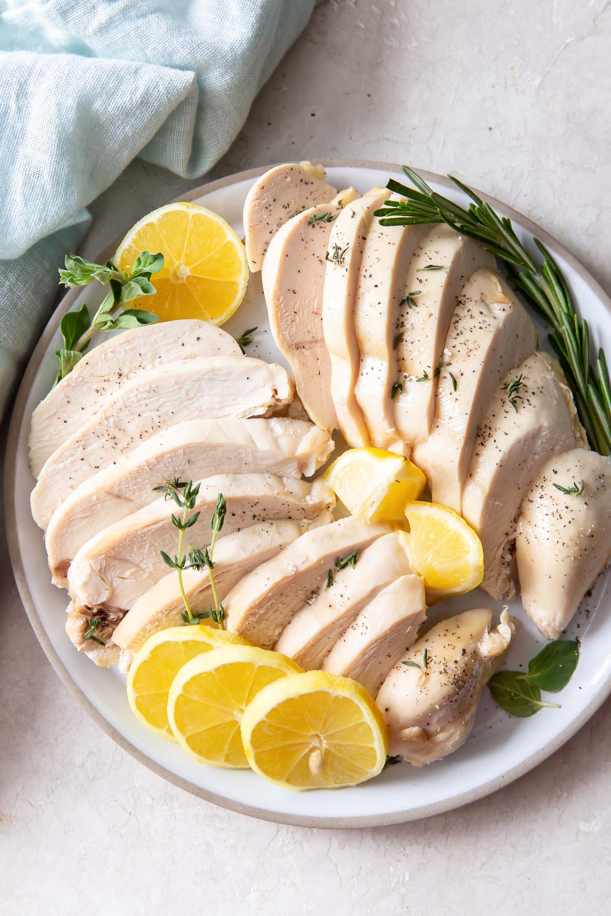 Sliced poached chicken on a plate with lemon and herbs.
