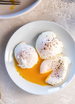 Three poached eggs on a plate with one egg sliced open so yolk runs out.