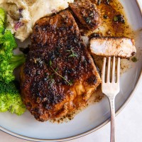Pan fried pork chop with bite on a fork served on a plate with mashed potatoes and broccoli.