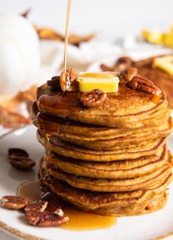 pouring syrup onto a stack of eight pumpkin pancakes with a pat of butter and pecan halves