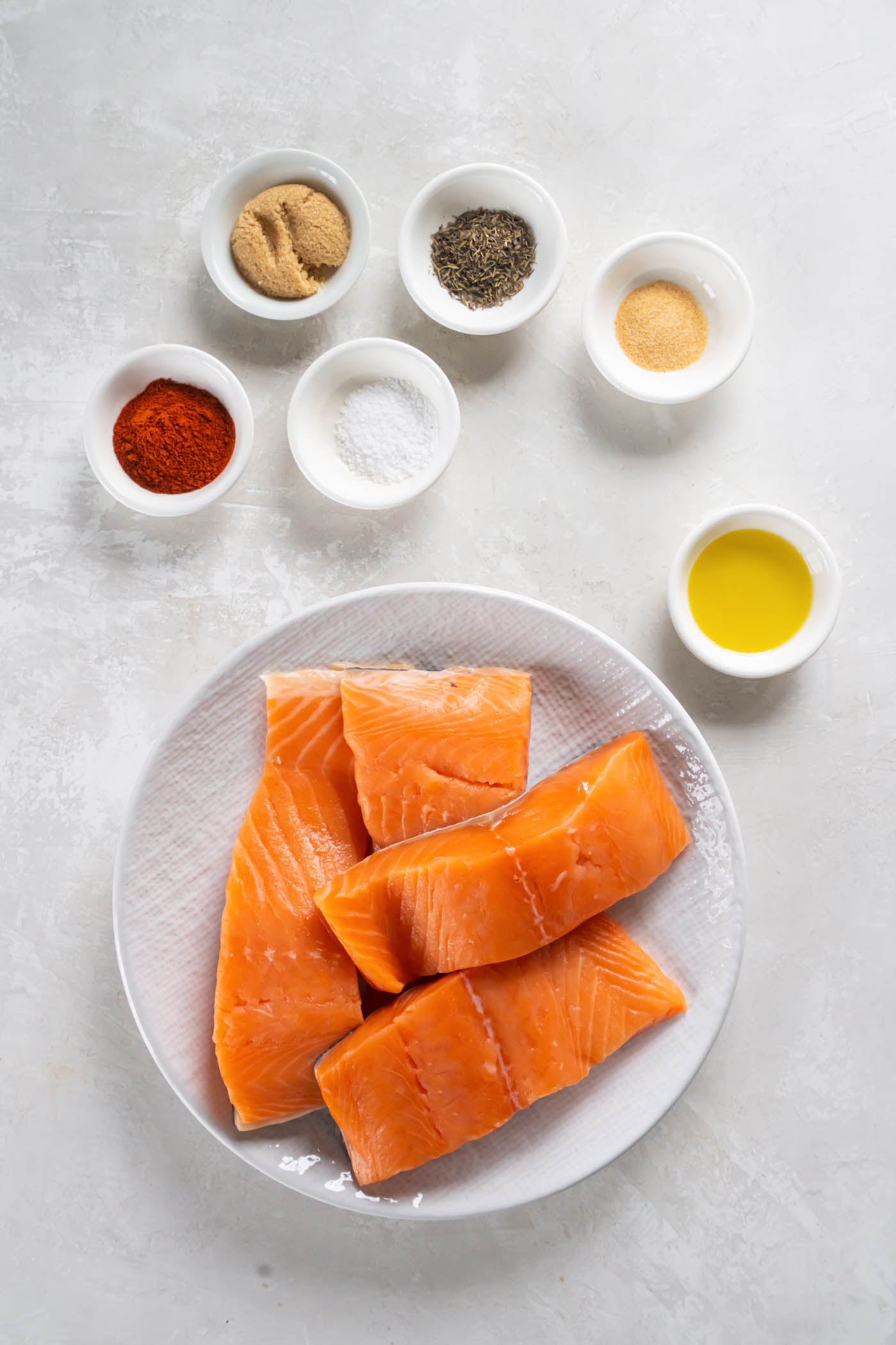 Spices and olive oil in small bowls, and four raw salmon fillets on a plate.