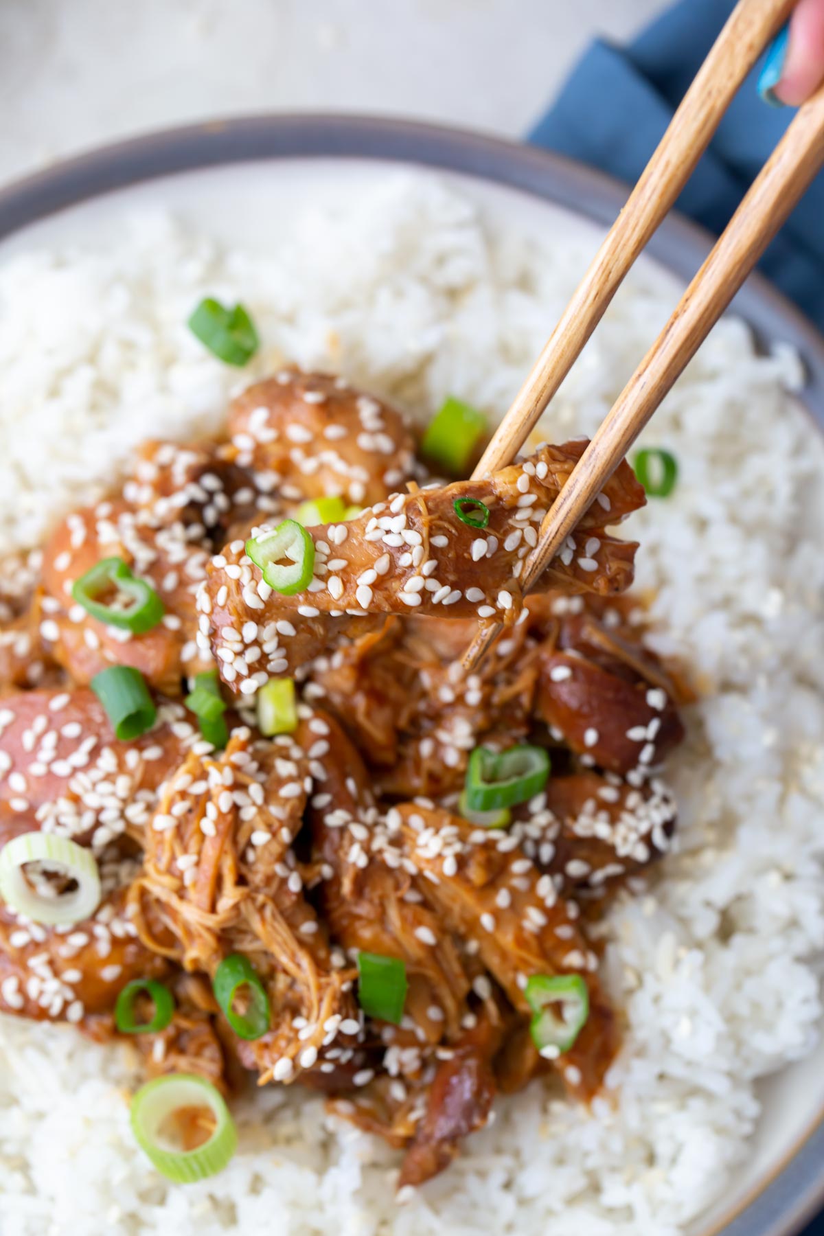 A bite of sesame chicken held with chopsticks over a plate of rice and sesame chicken.