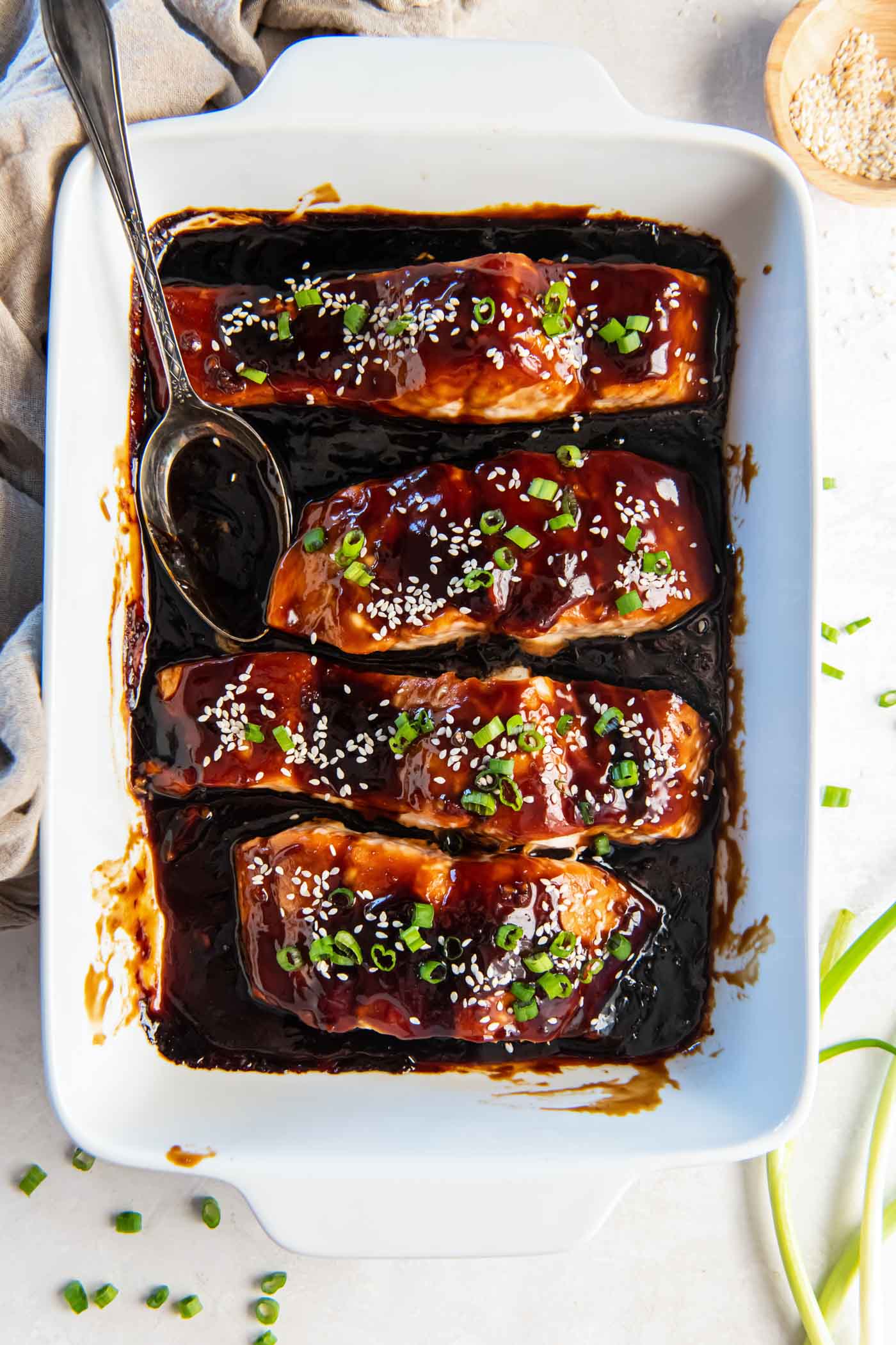 Baked teriyaki salmon in a white baking dish garnished with chopped green onions and sesame seeds.