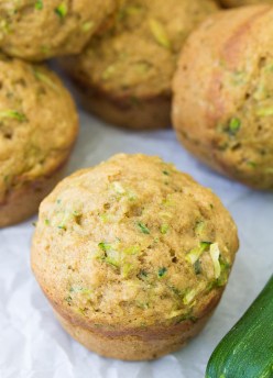 Close up of a zucchini muffin with a pile of muffins in the background and a fresh zucchini on the side.